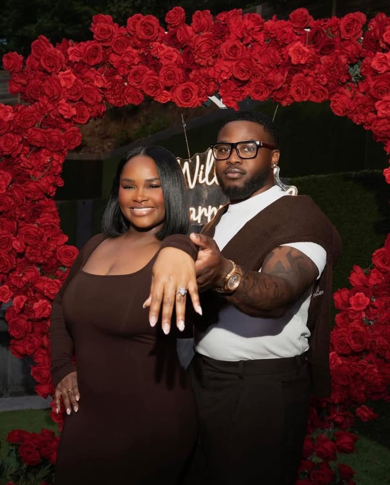 Couple photographed inside a red rose heart arch with a Will You Marry Me neon sign