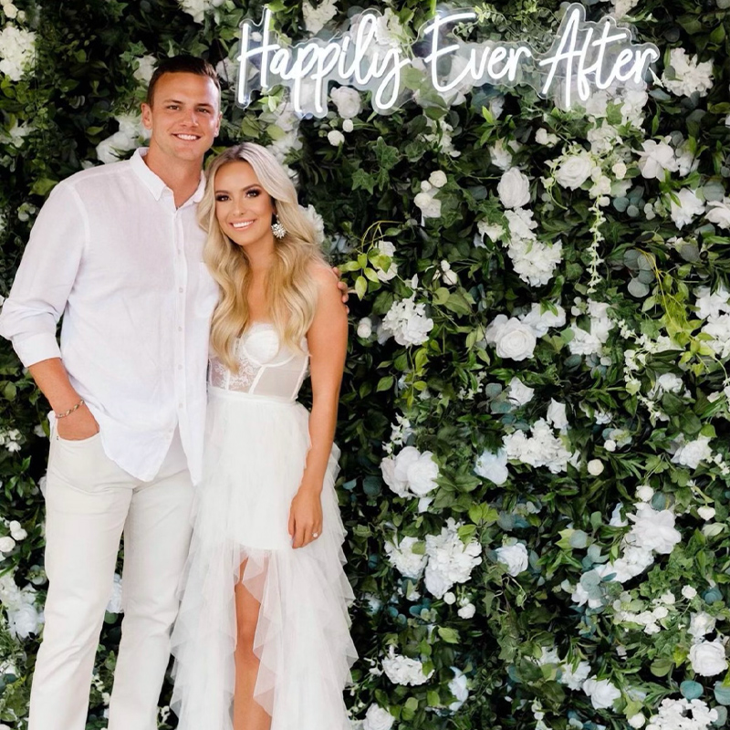Pro experience Flower wall rentals with green and white foliage with neon rental and couple posing for a picture.